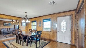 A dining area with a wooden table and chairs, a chandelier, and a view of the outdoors through windows. - Reel'em In Lodge - A Reel Fisherman Lodge! (St Bernard)