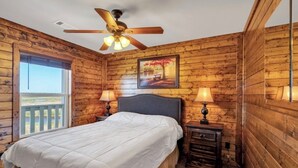 A bedroom with a wooden log wall, a bed with white bedding, two bedside lamps, a ceiling fan, and a window with a view. - Reel'em In Lodge - A Reel Fisherman Lodge! (St Bernard)