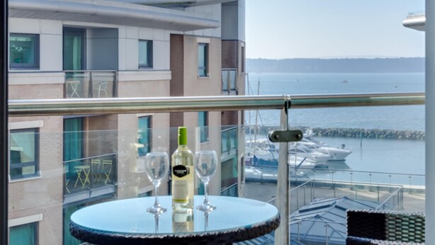A balcony with a round table, two wine glasses, and a bottle of wine. The view includes a body of water and boats.