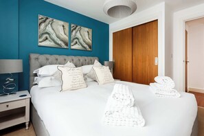 Cama bem arrumada com lençóis brancos, cabeceira cinza com capitonê e duas obras de arte abstratas na parede.