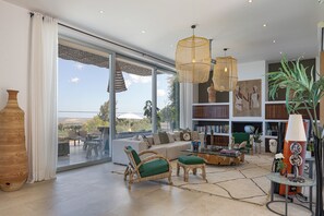 A spacious living room with a large sofa, wicker chairs, a glass coffee table, and a bookshelf filled with books. - Villa Amnalys - Sea View, Gardens & Spa by etouri (Rethymno)