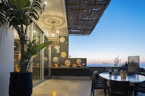 An outdoor dining area with a round table and chairs, surrounded by potted plants and decorative items. - Villa Amnalys - Sea View, Gardens & Spa by etouri (Rethymno)