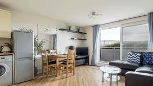 Um apartamento moderno com cozinha, sala de jantar e sala de estar. Possui máquina de lavar, mesa de jantar com cadeiras, televisão, sofá e varanda com vista.