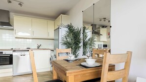Uma cozinha compacta com armários brancos, lava-louças e micro-ondas. Há uma mesa de jantar com cadeiras e um vaso de planta.