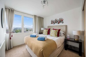 A hotel room with a large bed, two bedside tables, a nightstand, a view of the city, and a decorative wall piece.