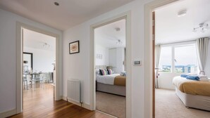 A modern hotel room with a wooden floor, a bed with a beige blanket, a large window with curtains, and a view of the city. - Tides - Poole Town Views, On-site Parking (Poole)