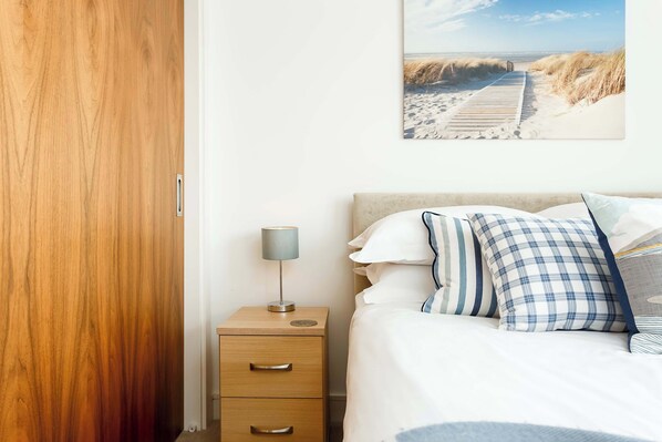 Una camera da letto con un armadio in legno, un comodino con una lampada, un letto con biancheria da letto bianca e cuscini a quadri, e un quadro a tema spiaggia.