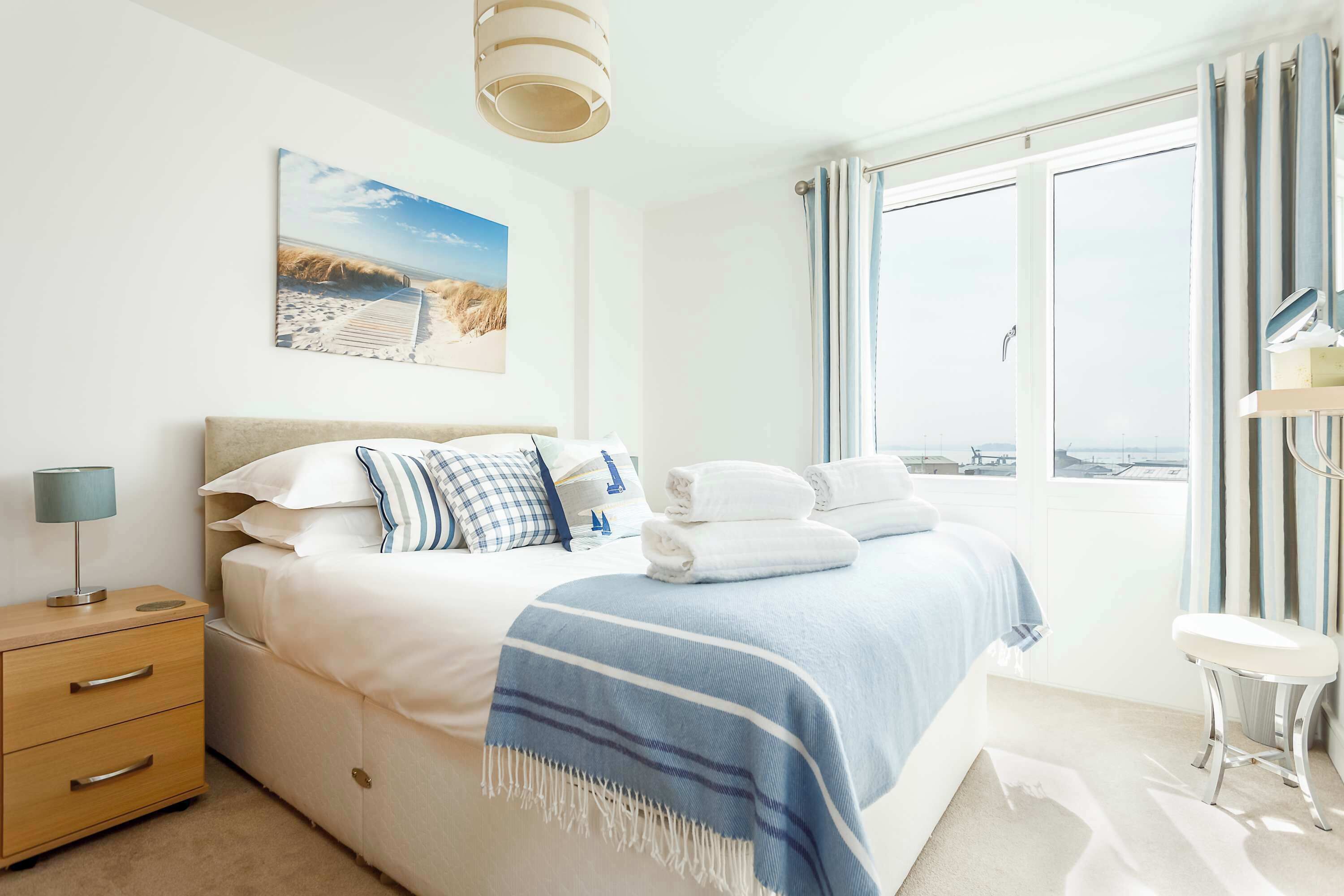 A bedroom with a large bed, a nightstand, a lamp, a view of the beach, and a window with curtains.