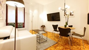 A modern living room with a white sofa, a dining table with chairs, a TV mounted on the wall, and a floor lamp. - Malasaña Downtown (Madrid)