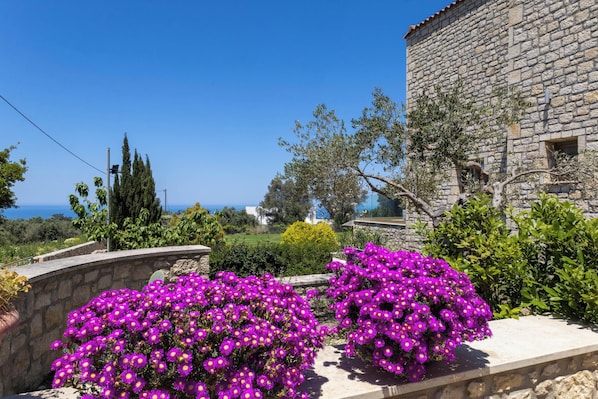 A stone building with a clear view of the sea, surrounded by lush greenery and vibrant purple flowers.