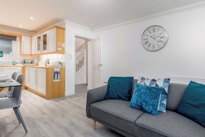 Una sala de estar moderna con un sofá gris y cojines verde azulado, una cocina con gabinetes blancos y un reloj de pared.