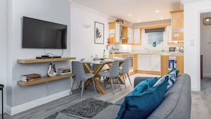 A modern living room with a flat-screen TV, a dining area with a wooden table and chairs, and a kitchen with white cabinets and a window. - Magnolia House - Townhouse in Old Town Poole (Poole)