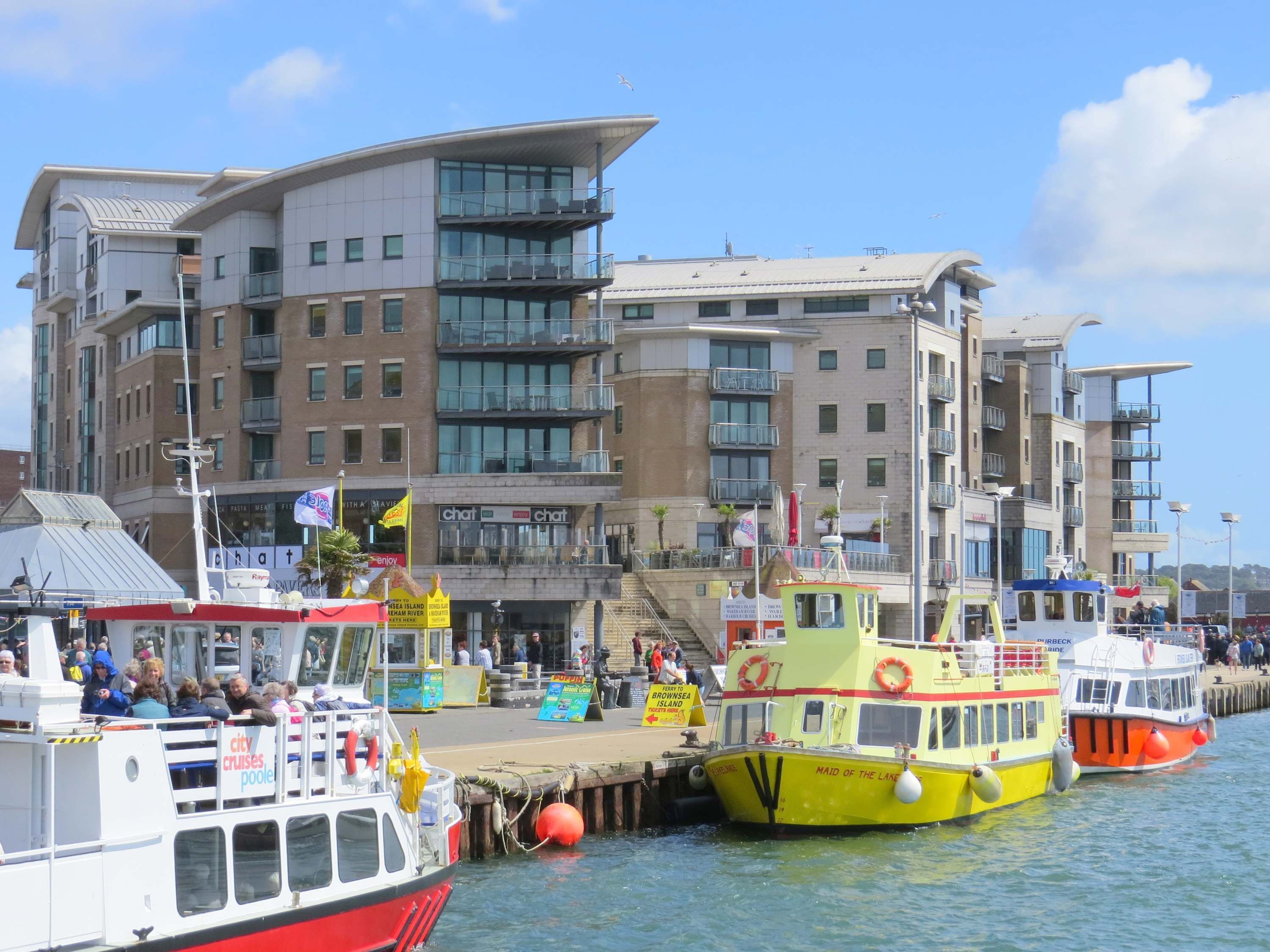A waterfront with several boats docked, a modern building with balconies, and a clear sky.