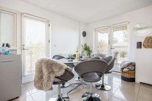 Una cocina moderna con armarios blancos, un refrigerador blanco, una mesa de comedor con sillas y una ventana con persianas.