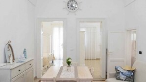 A room with a dining table, chairs, a clock on the wall, and a sofa with a patterned pillow. - WePuglia - Domus Neptuni (Monopoli)