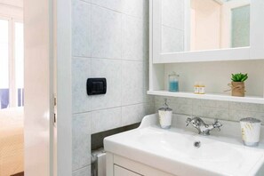 A bathroom with a white sink, a mirror, and a shelf with toiletries. - WePuglia - Domus Neptuni (Monopoli)
