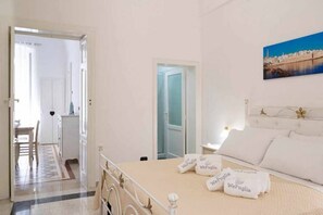 A bedroom with a bed, a nightstand, a painting on the wall, and a door leading to another room. - WePuglia - Domus Neptuni (Monopoli)
