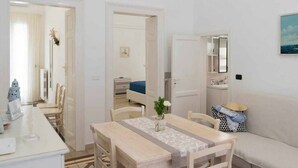 A compact room with a dining table, chairs, a sofa, a small kitchen area, and a door leading to another room. - WePuglia - Domus Neptuni (Monopoli)