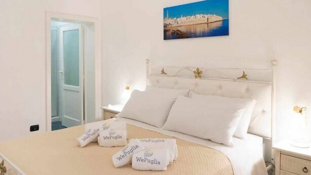 A hotel room with a bed, a nightstand, a bedside table, a framed picture on the wall, and a door leading to another room. - WePuglia - Domus Neptuni (Monopoli)
