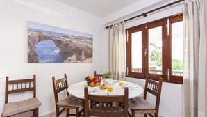 Un coin repas avec une table ronde, quatre chaises en bois, un panier de fruits et un tableau représentant un paysage côtier.
