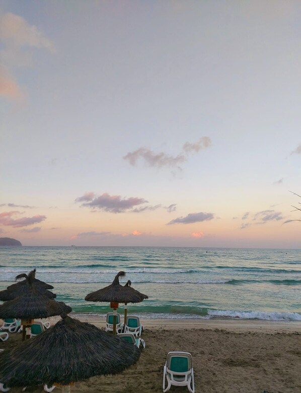 Una playa con sombrillas de techo de paja y sillas blancas de plástico.