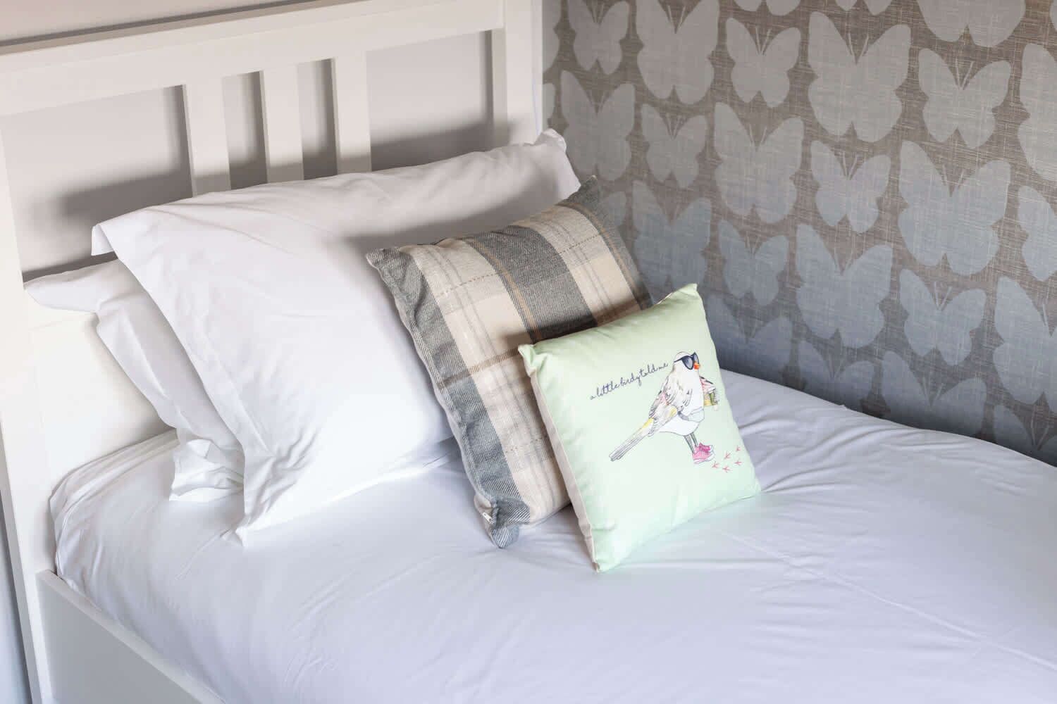 Un lit avec une literie blanche, un coussin à carreaux et un coussin orné d’une illustration d’oiseau.