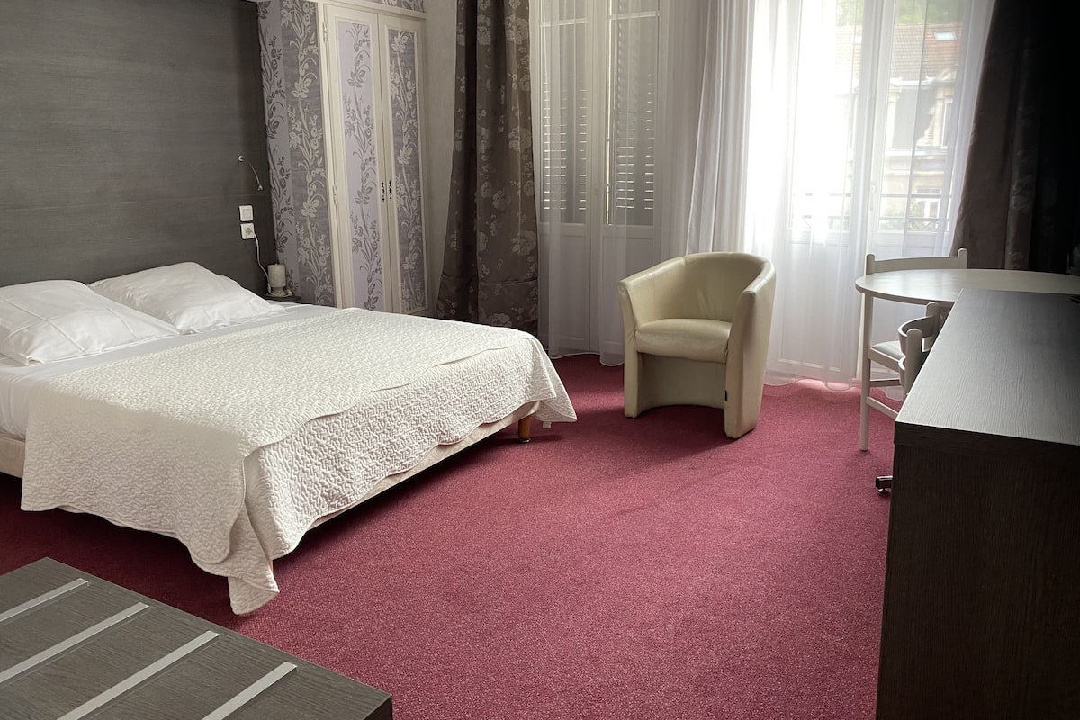 A hotel room with a bed, a chair, a small table, and a window with curtains.