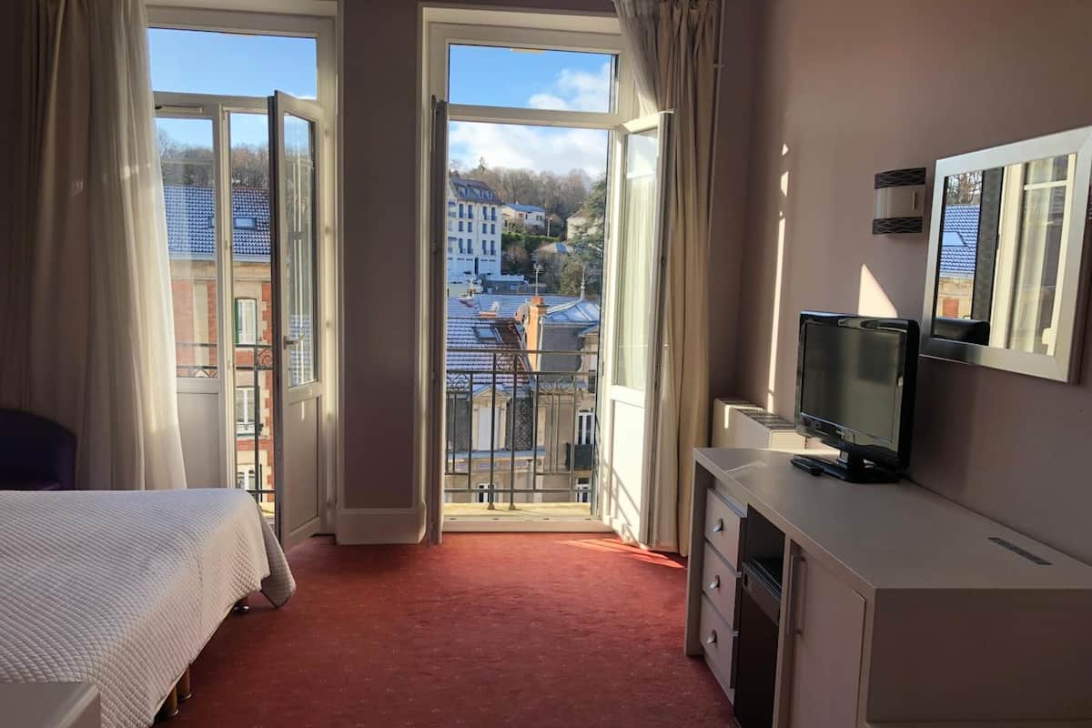A hotel room with a bed, a desk with a TV, and a balcony with a city view.
