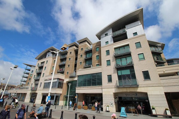 A multi-story building with balconies, a café named "Quay Café," and a pedestrian area with people walking and a cyclist. - Marina Secret - with Free Parking (Poole)