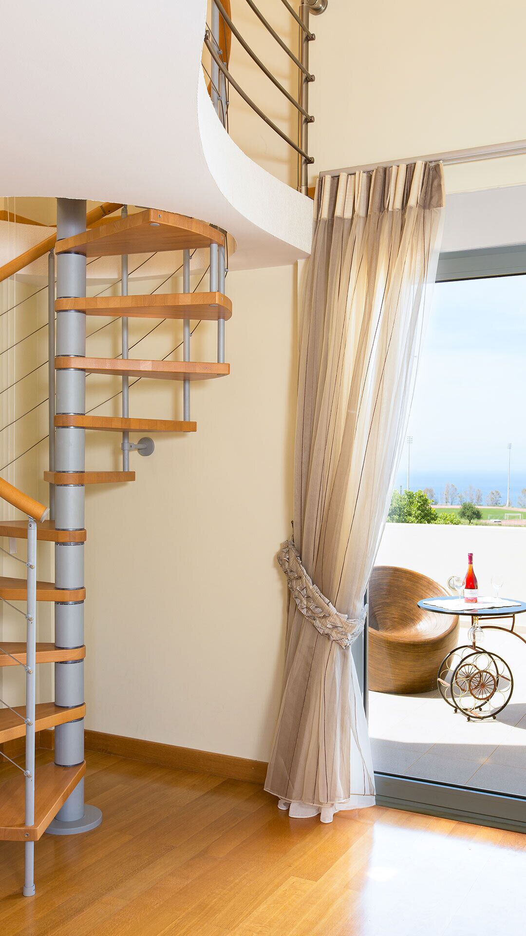 A modern interior with a spiral staircase, a large window with a view, and a round table with a bottle and glass.