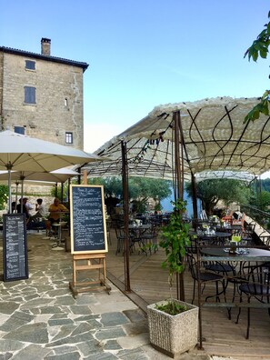 An outdoor café with stone flooring, tables, and chairs, a chalkboard menu, and a building in the background.