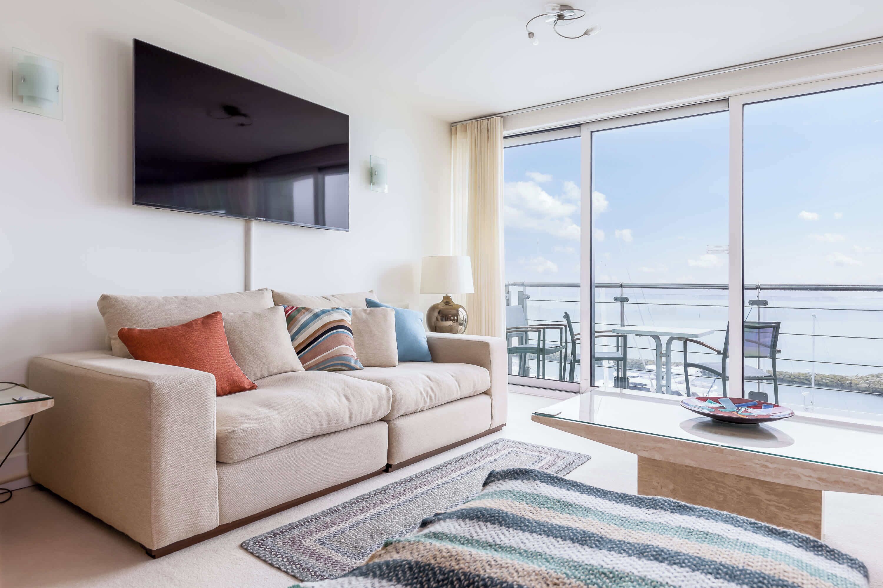 Un salon moderne avec un canapé, un téléviseur à écran plat, une table basse en verre et une vue sur la mer grâce à de grandes fenêtres.