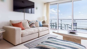 A modern living room with a sofa, a flat-screen TV, a glass coffee table, and a view of the sea through large windows. - Riva -180-Degree Panoramic Views from Balcony (Poole)