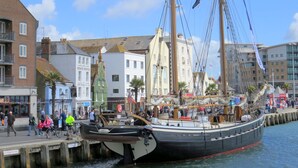 Ein historisches Segelschiff liegt an einem Kai, im Hintergrund sind Gebäude und Fußgänger zu sehen.