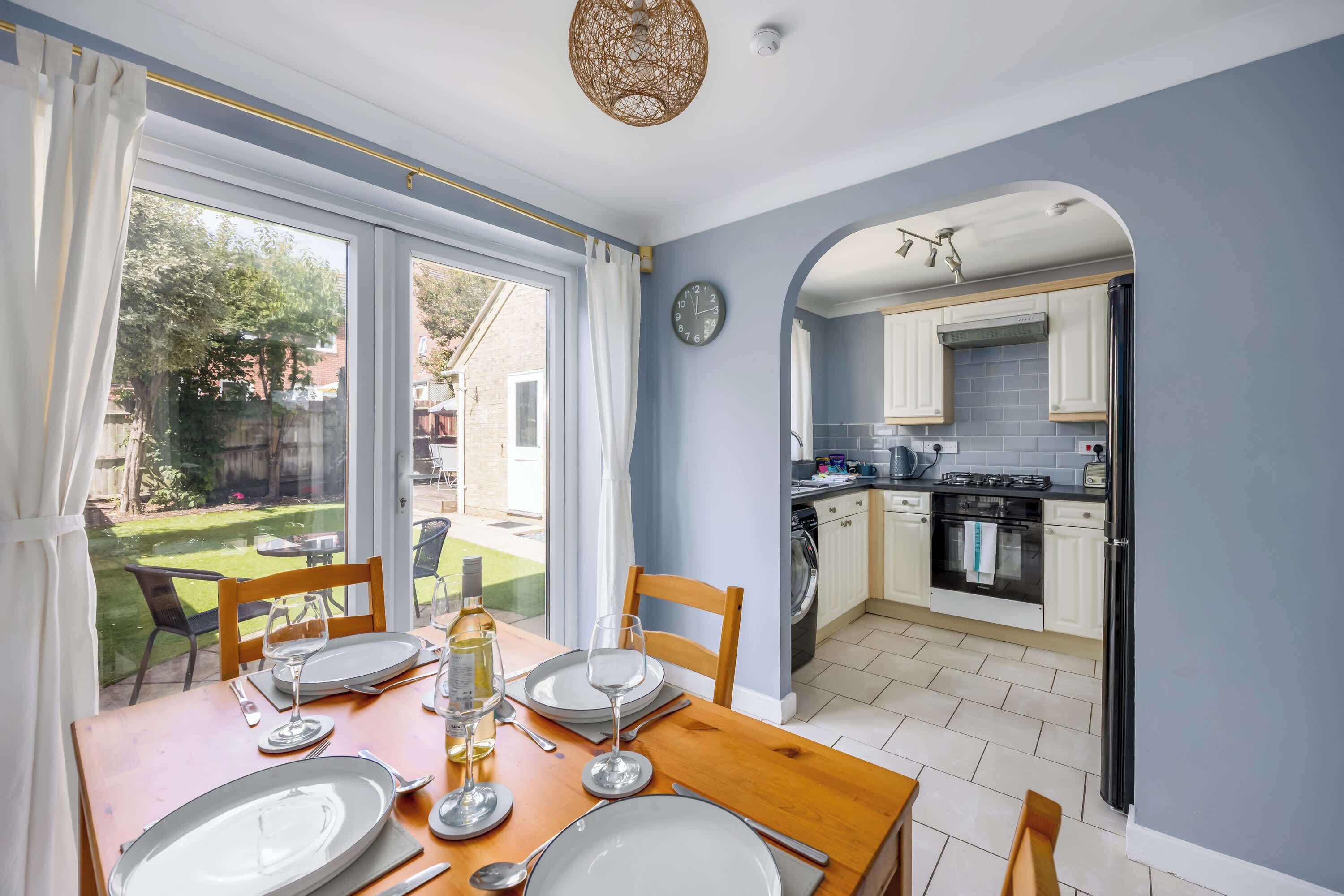 A dining area with a table set for four, a kitchen with white cabinets, and a view of the garden through the window.
