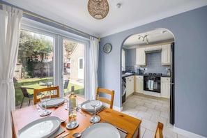 A dining area with a table set for four, a kitchen with white cabinets, and a view of the garden through the window. - Palma - Coastal Home near Harbour and beach (Poole)