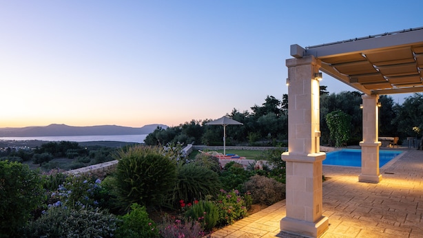 Ein überdachter Außenbereich mit Pool, Steinsäulen und Blick auf das Meer im Abendlicht.