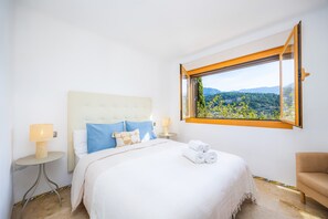 A hotel room with a bed, a chair, a nightstand, a lamp, and a window with a view of greenery and mountains. - Renovated 2026! Air-Conditioned Apartment with Seaview, shared pool and Wi-Fi in Port de Sóller (Port de Soller)