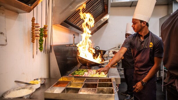 A chef cooking with a large wok over an open flame in a professional kitchen. - Hotel Indian Heritage (Raipur)