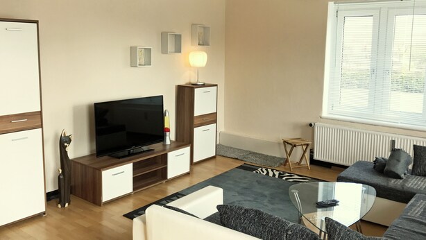 Ein modernes Wohnzimmer mit Flachbildfernseher, Holzschränken, einem Glastisch und einem grauen Sofa.