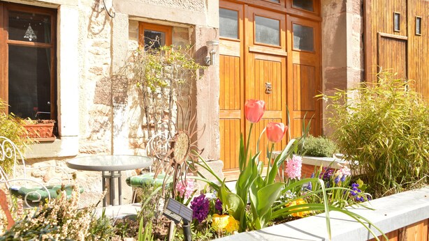 A stone building with a wooden door and windows, a garden with various plants and flowers, and a small table and chair outside. - Small rustic cottage with terrace u. Hochgarten in outskirts (Tignécourt)