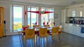 Ett modernt matområde med utsikt över havet, ett kök med vita skåp och ett matbord dukat med röda servetter.