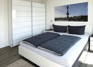 Une chambre moderne avec un grand lit, une table de chevet, un téléviseur fixé au mur et un tableau représentant un phare.