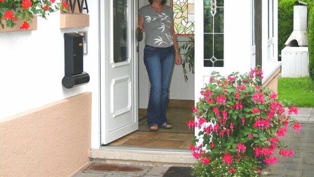 A woman standing at the entrance of a house with a red tiled roof, white walls, and a glass door. - Apartment 1 - House Erna (Bad Wildungen)