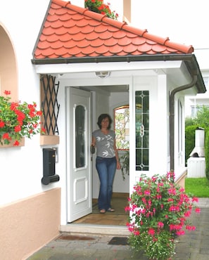 Une femme se tient à l’entrée d’une maison dont le toit est recouvert de tuiles rouges, les murs sont blancs et la porte est en verre.