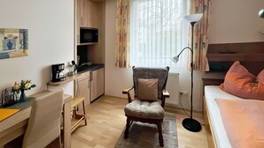 A compact hotel room with a kitchenette, a dining area, a comfortable armchair, a bed with pink pillows, and a large window with curtains. - Apartment 1 - House Erna (Bad Wildungen)