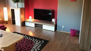 Ett modernt vardagsrum med en platt-TV, en röd accentvägg, en vit vas med blommor, en golvlampa och en mönstrad matta.