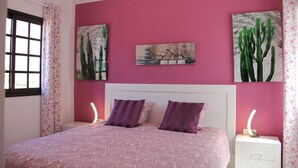 Une chambre avec un mur rose, un lit recouvert d’une couvre-lit à motifs, deux oreillers, une table de chevet avec une lampe et deux œuvres d’art mural représentant des cactus.