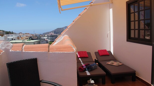 Un balcon avec vue sur la ville, deux chaises longues avec des coussins rouges, une chaise et une table sur laquelle se trouvent une serviette et un objet enroulé.