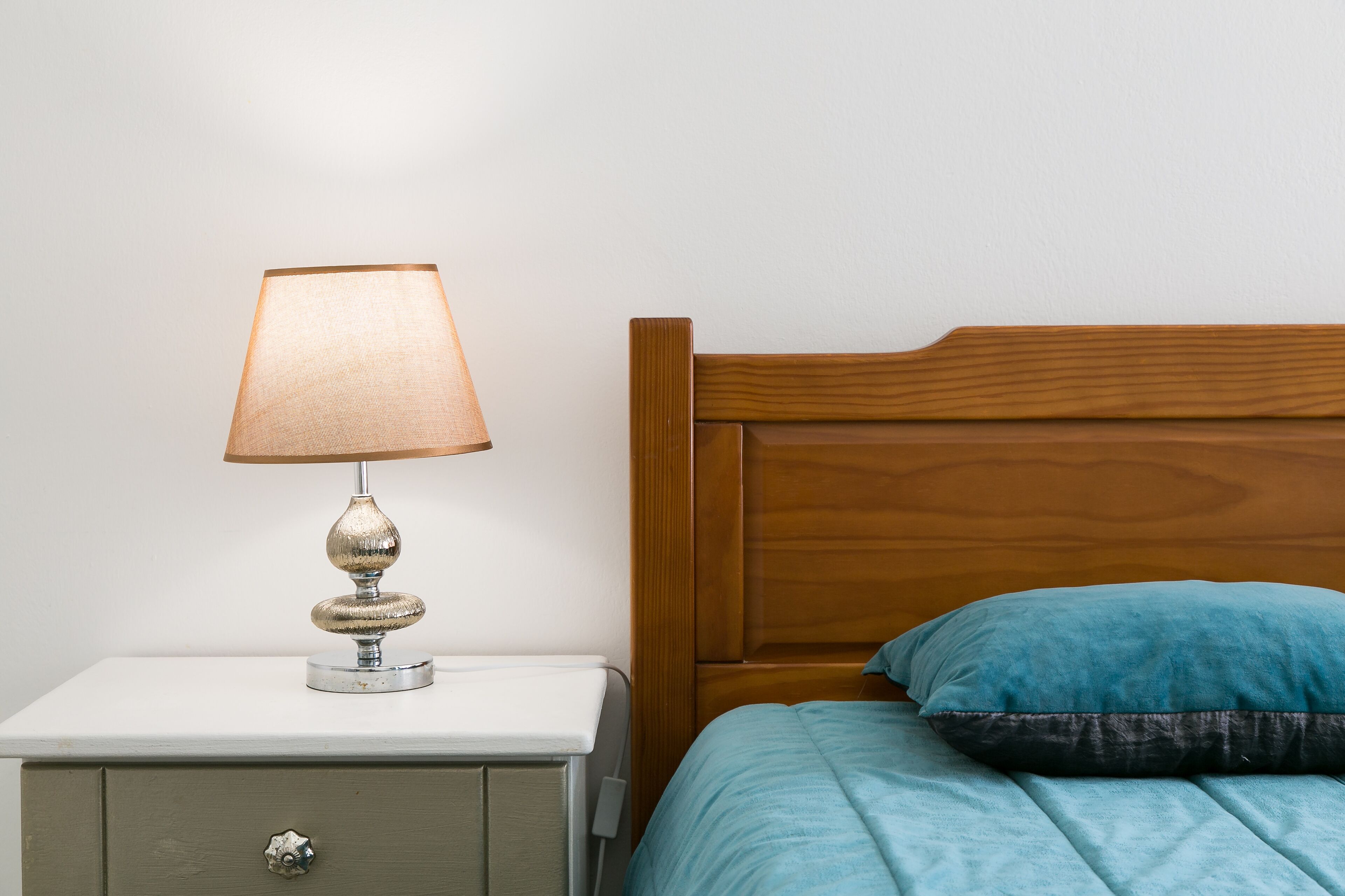 Un lit avec une tête de lit en bois, une table de chevet blanche avec une lampe et un seul tiroir.